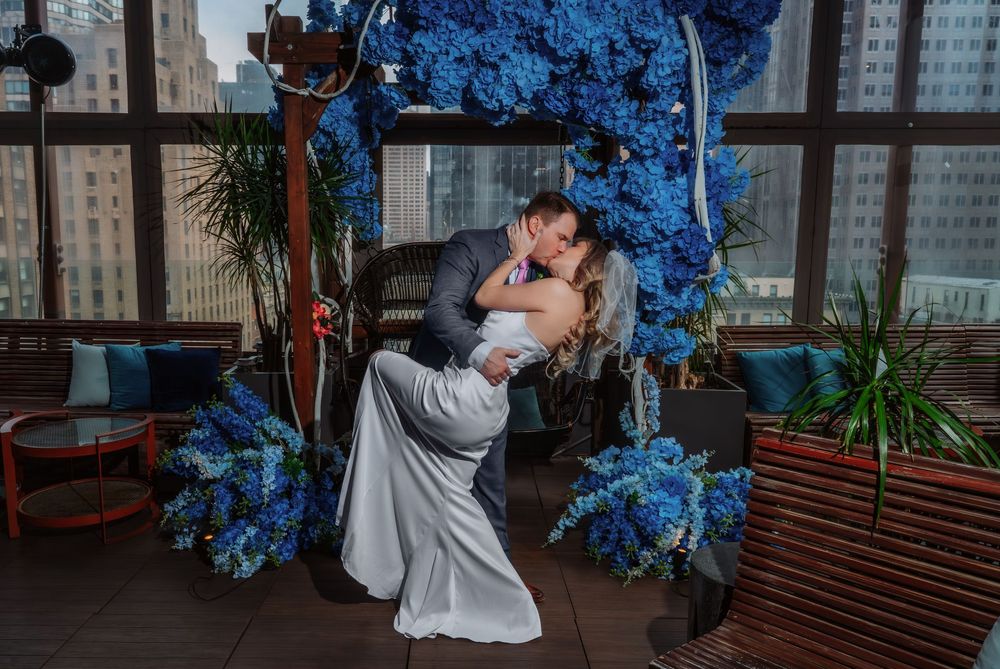 A couple embraces under an arch decorated with vibrant blue flowers in an urban setting.