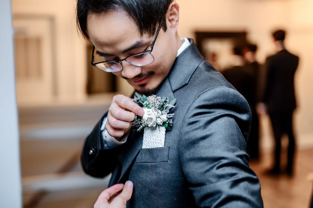 A groom in a charcoal suit and bow tie adjusts his boutonniere during an outdoor wedding ceremony.