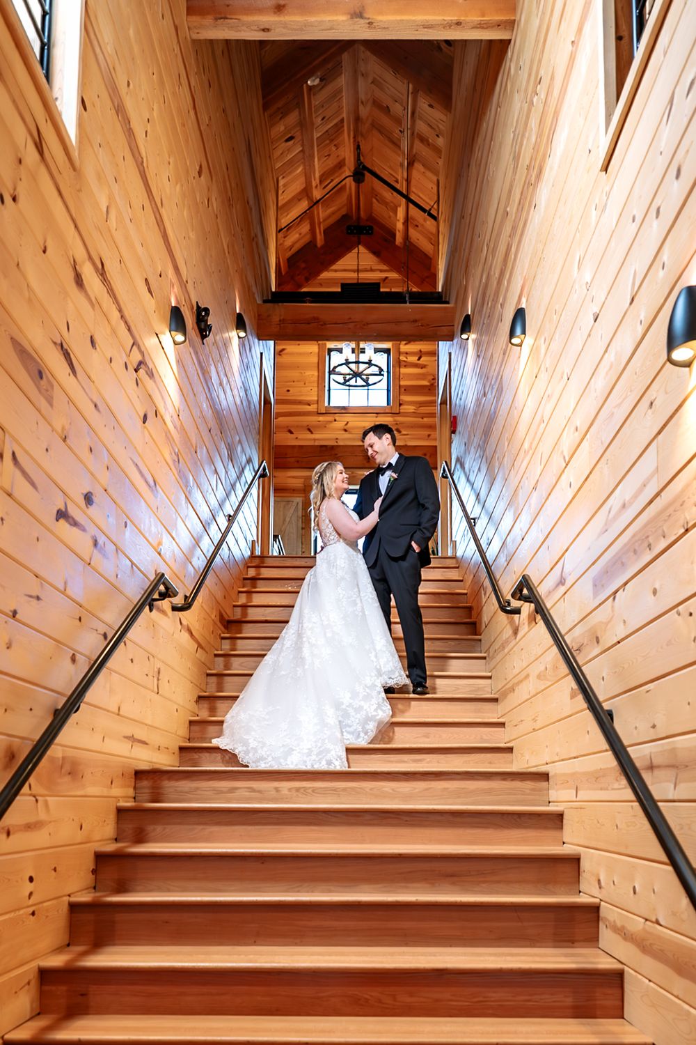 Bride and groom embrace on wooden staircase in rustic chapel venue, bride in lace gown, groom in black suit.
