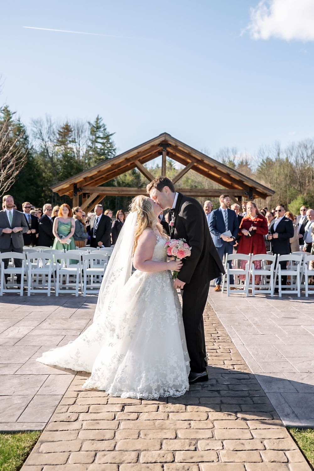 Bride and groom share first kiss at outdoor wedding ceremony on stone patio with white chairs and wooden pavilion backdrop.