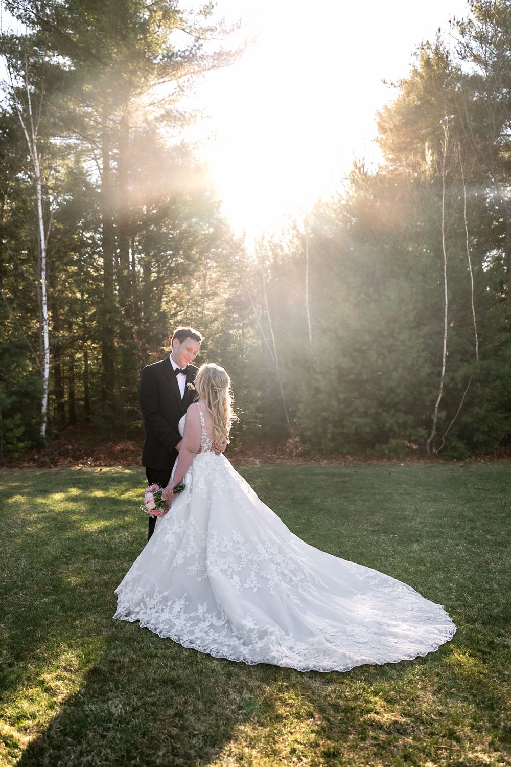 Bride in lace gown and groom in tuxedo share romantic moment on sunlit lawn surrounded by tall trees.