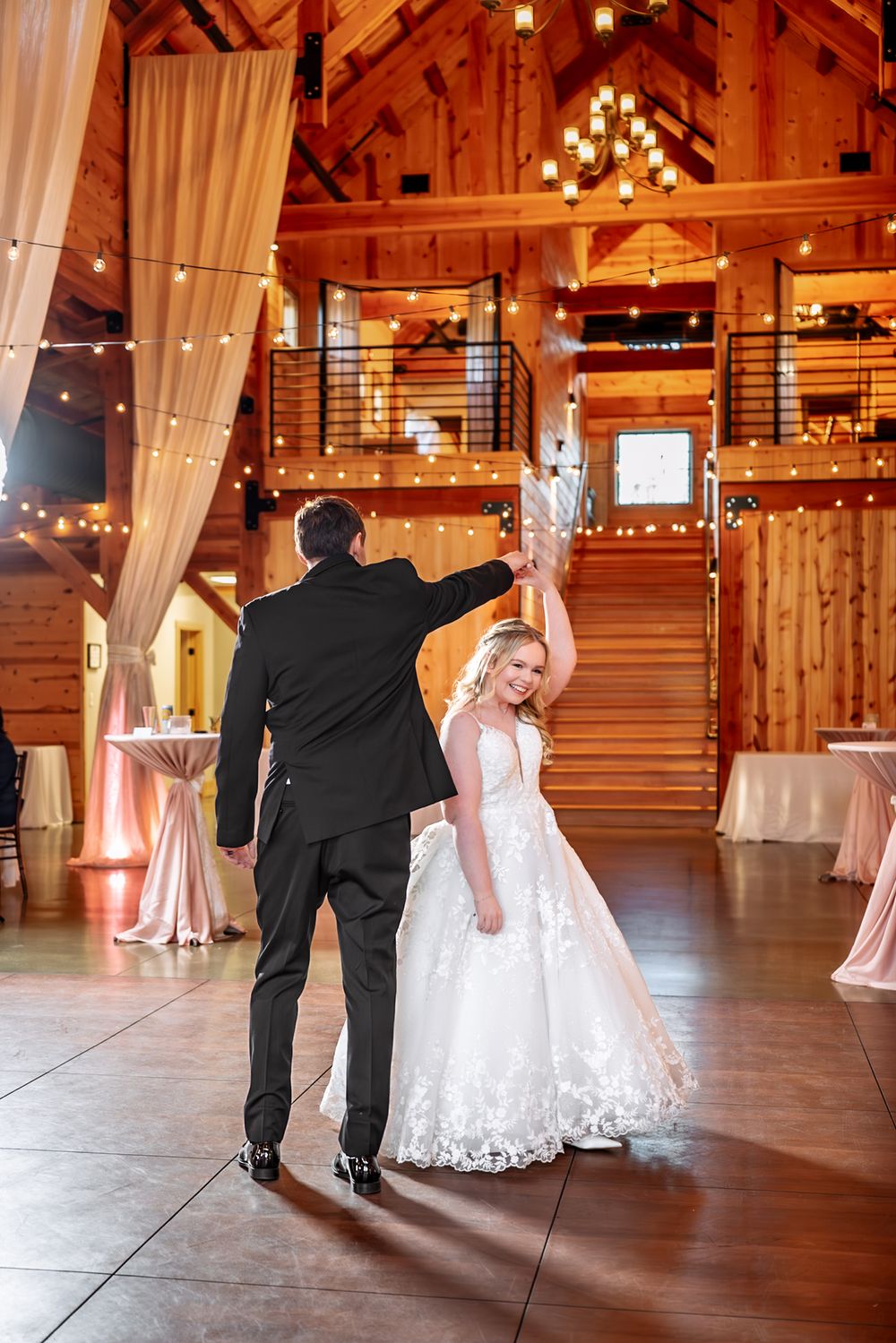 Bride and groom share first dance in rustic barn wedding venue with string lights and elegant draping.