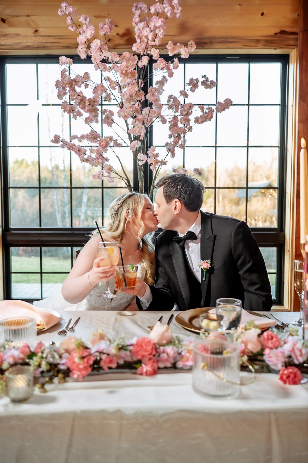 Bride and groom kissing at sweetheart table with pink cherry blossom centerpiece and floral arrangements at wedding reception.
