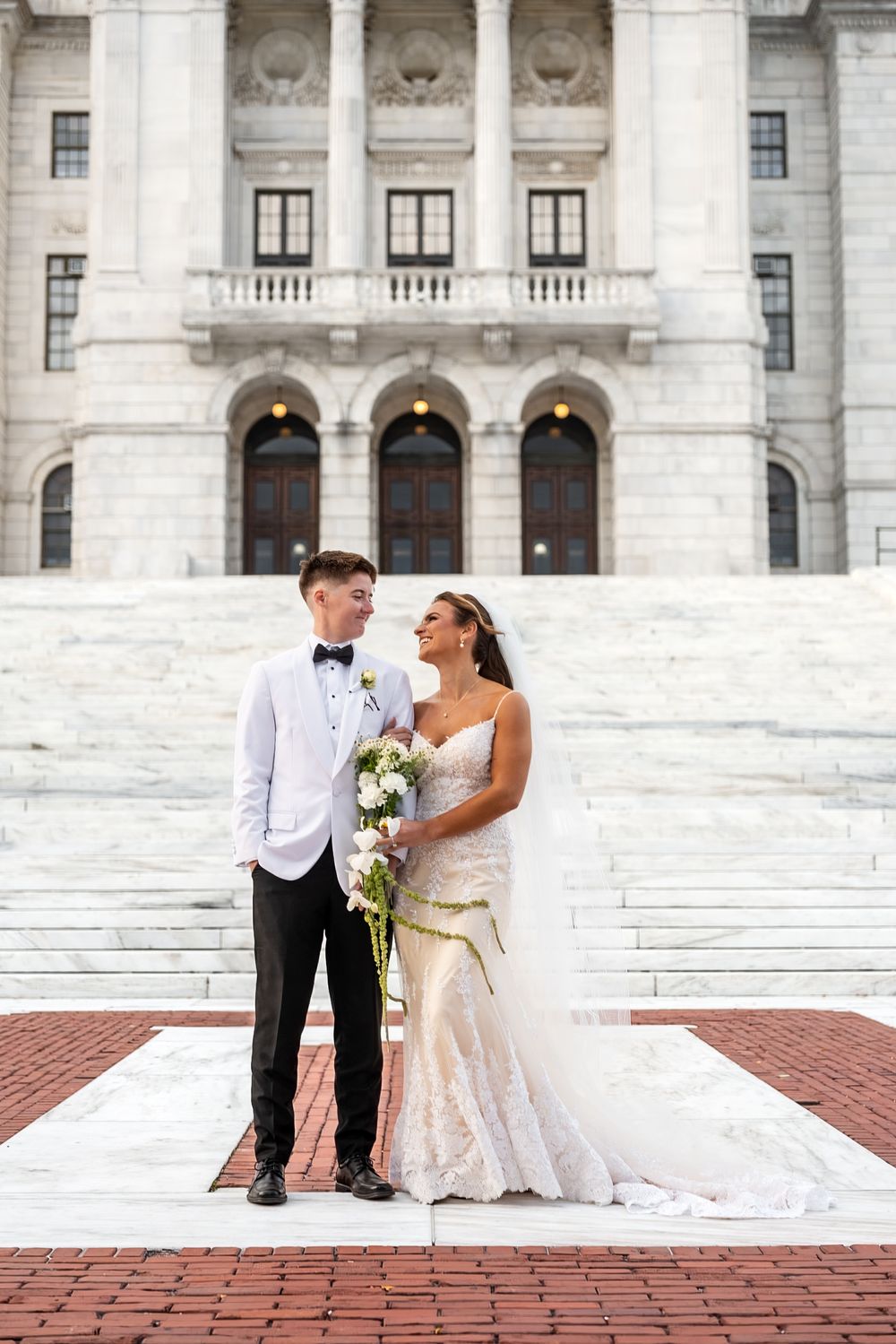 Newlywed couple sitting on marble steps of grand government building in formal wedding attire.