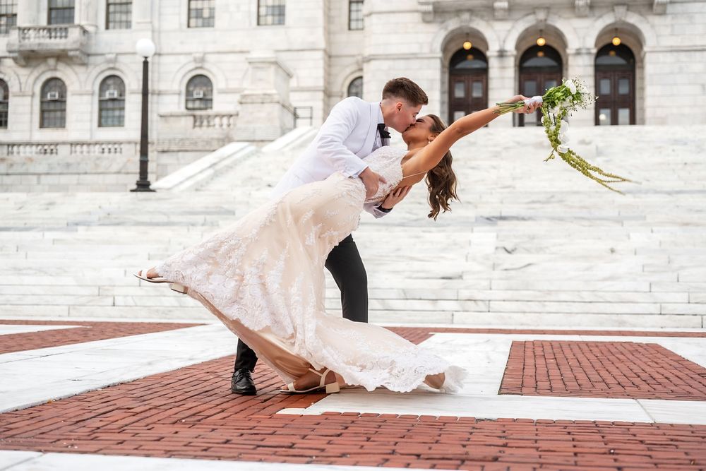 A romantic dip pose between newlyweds in formal attire on the steps of a grand stone building.