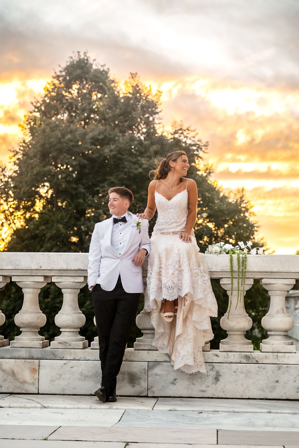 A couple in wedding attire sits on a stone balustrade at sunset with dramatic orange clouds in the background.