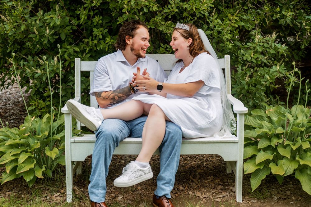 A couple sits together on a white garden bench surrounded by lush greenery.