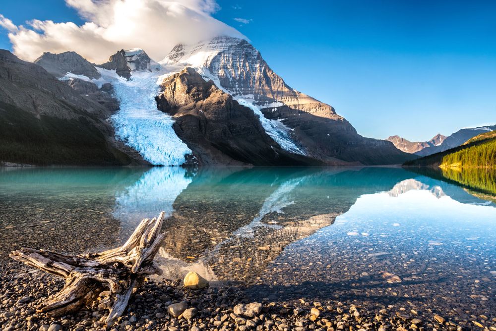 Mountain lake reflecting snow-capped peaks and glaciers with driftwood on rocky shore.