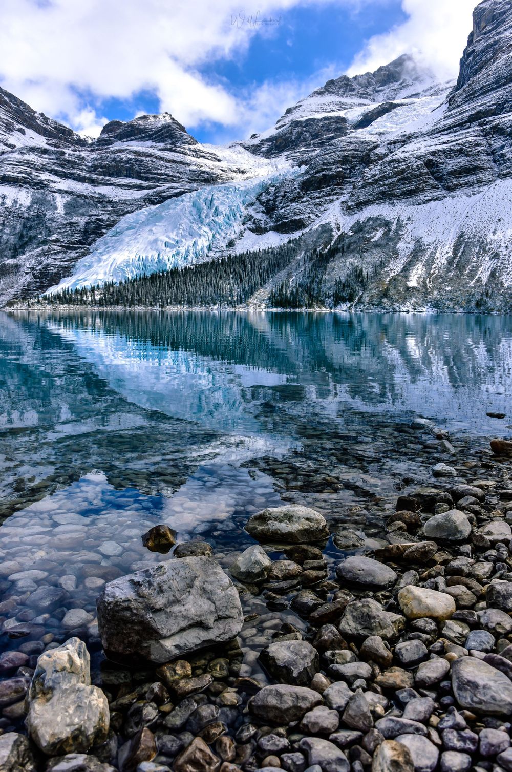 Glacial lake reflecting snow-capped mountains with rocky shoreline in Canadian Rockies.