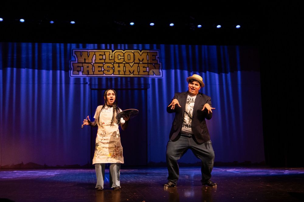 Two performers dance on stage beneath a Welcome Freshmen banner in a school-themed theatrical production.