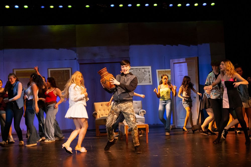 Energetic ensemble cast dances on stage during a lively musical theater performance with blue set design.