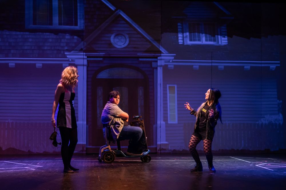 Three actors perform a dramatic scene outside a house backdrop under moody purple stage lighting.