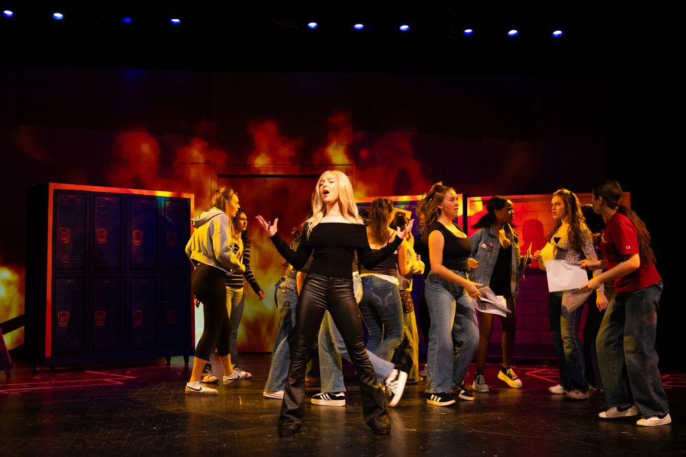 Performers on stage during a theatrical production with colorful lighting and locker backdrop.