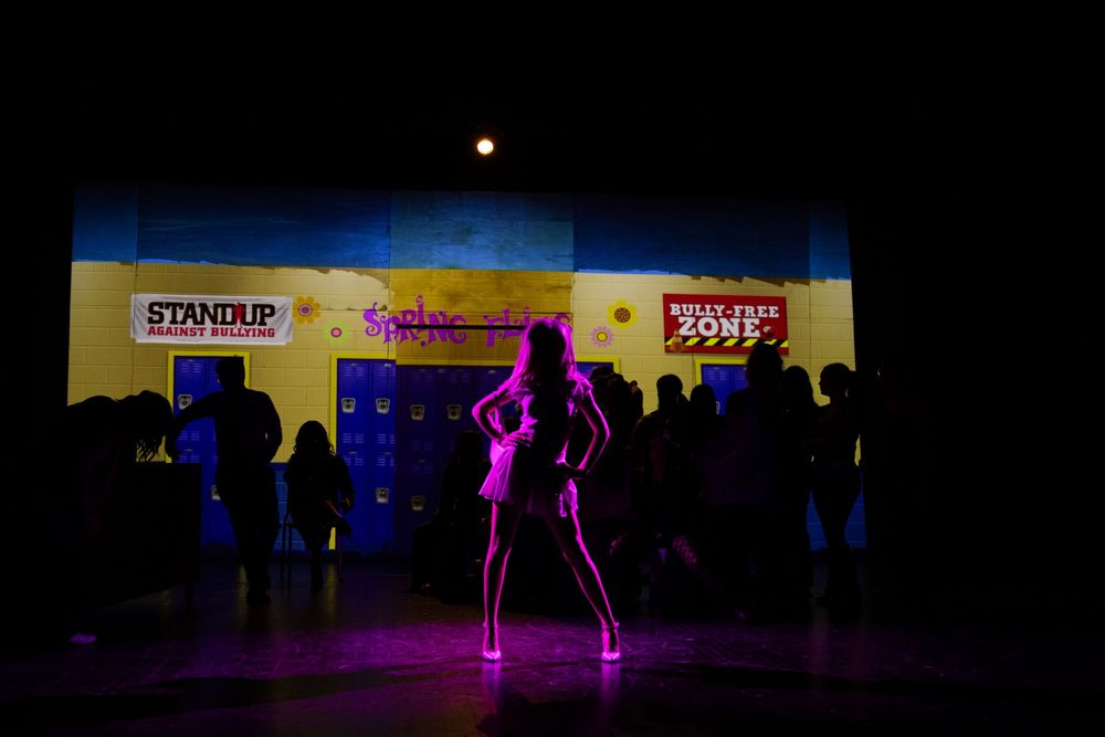 A solo performer stands dramatically in pink neon lighting on a darkened theater stage.