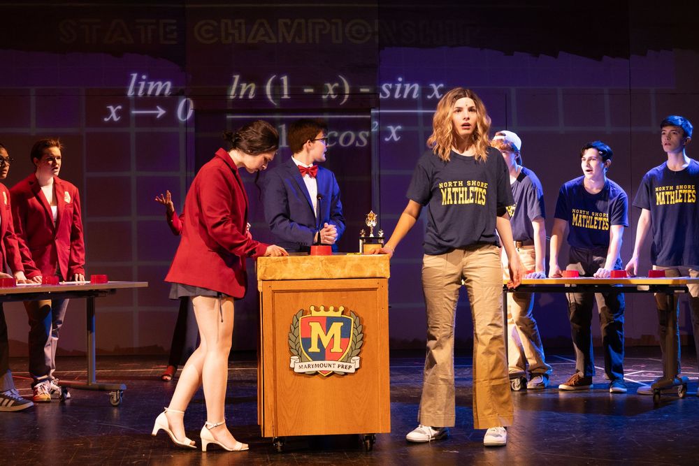 Students perform a school play on stage with math equations displayed in the background.
