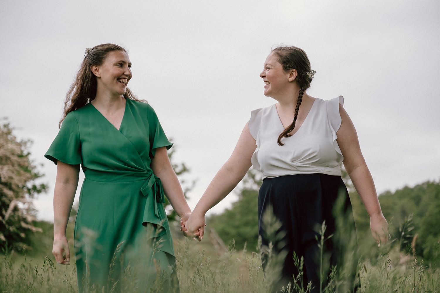 Two women in dresses hold hands and smile at each other while walking through a green field.