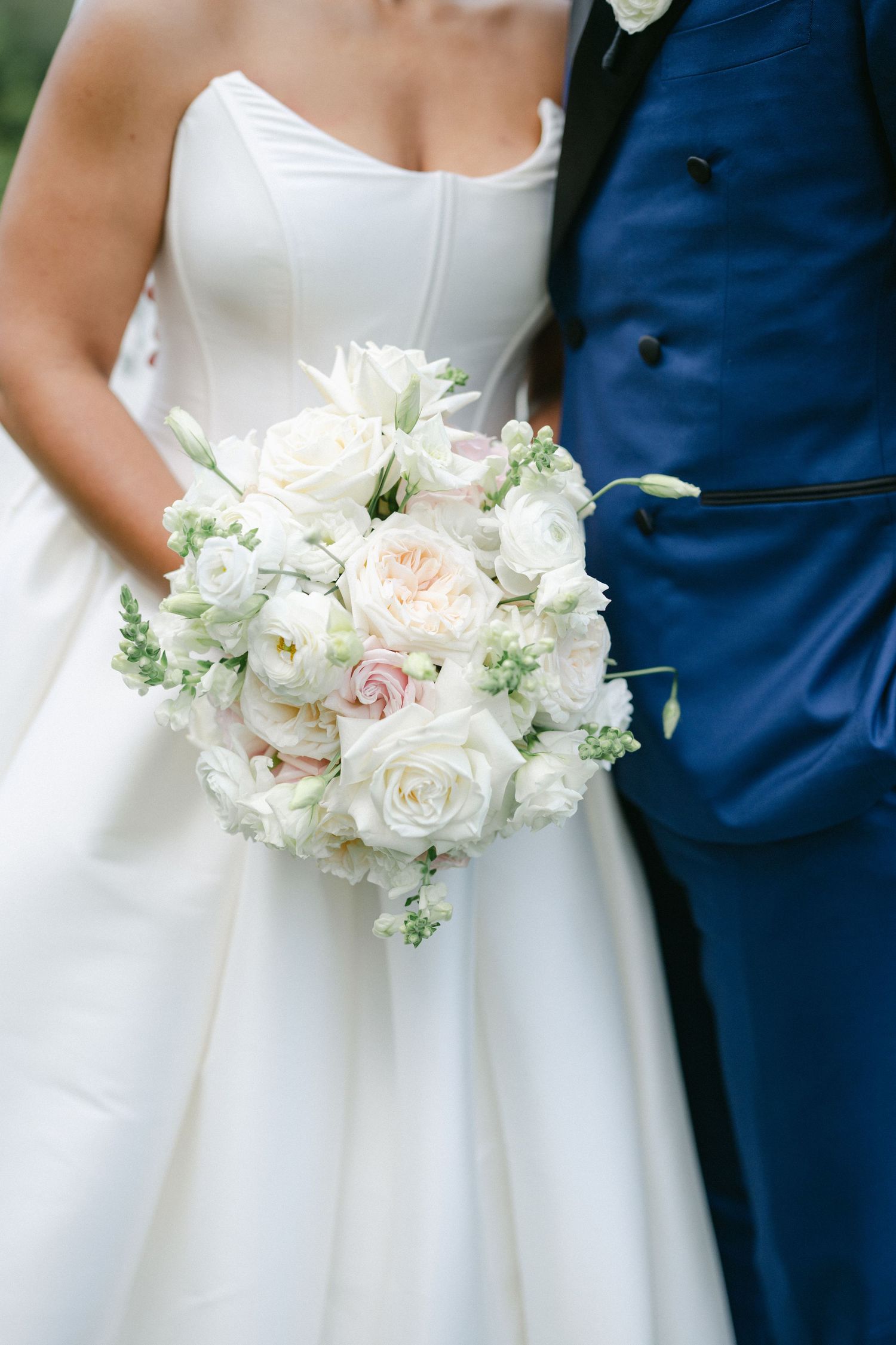 Close-up detail of white and blush pink bridal bouquet against navy suit and white wedding dress.