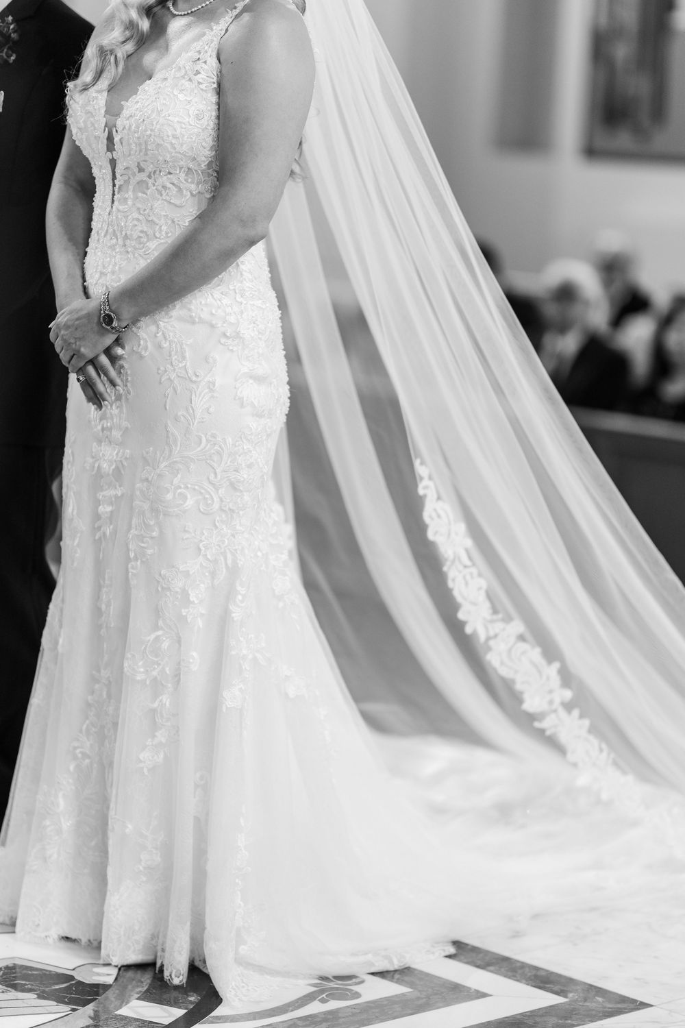 Close-up details of a white lace wedding dress with flowing veil trailing behind on a patterned floor.