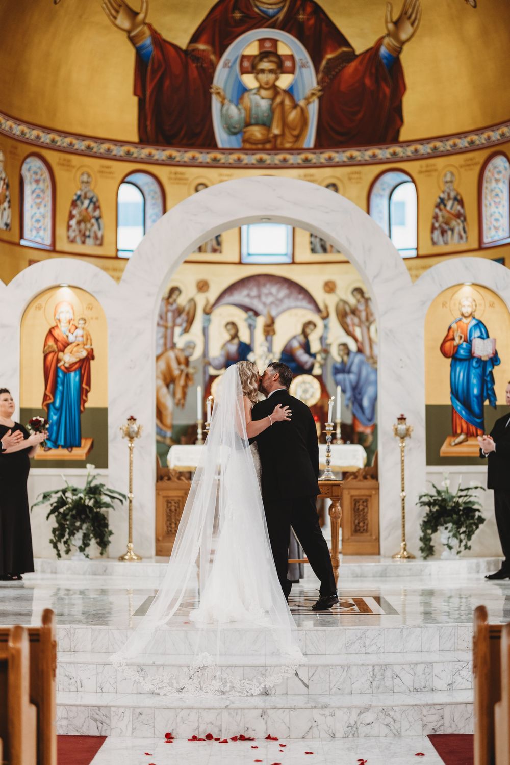 Wedding ceremony taking place in an ornate Orthodox church with golden walls and religious murals.