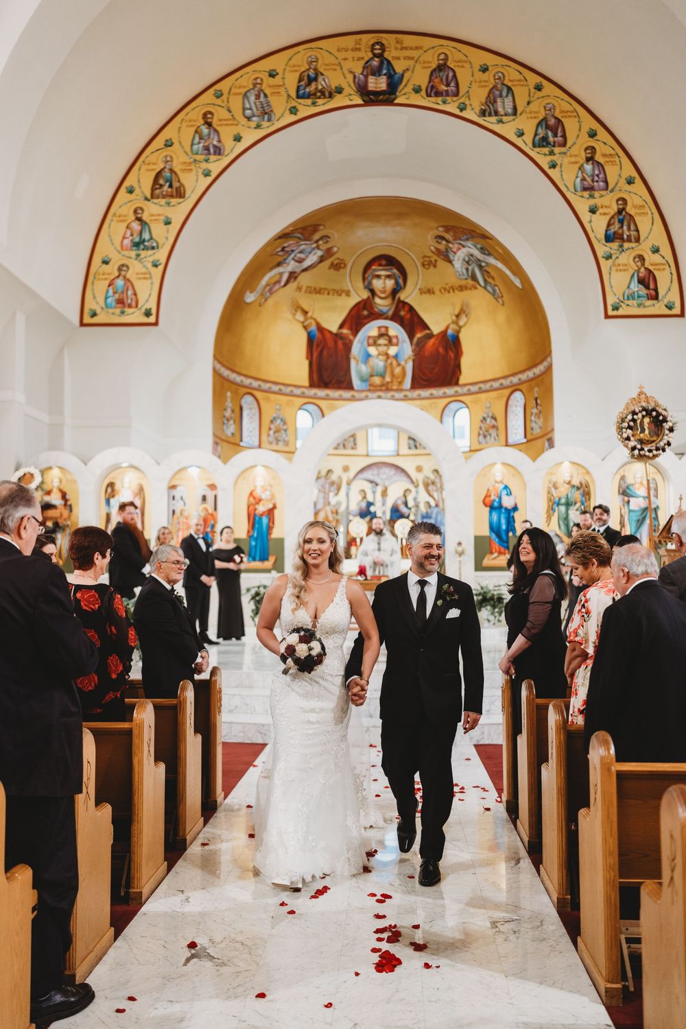 Wedding processional in Orthodox church with guests lining wooden pews and rose petals scattered on aisle.