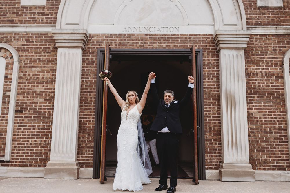 Wedding party members celebrate with raised arms outside a classic brick church building with white columns.