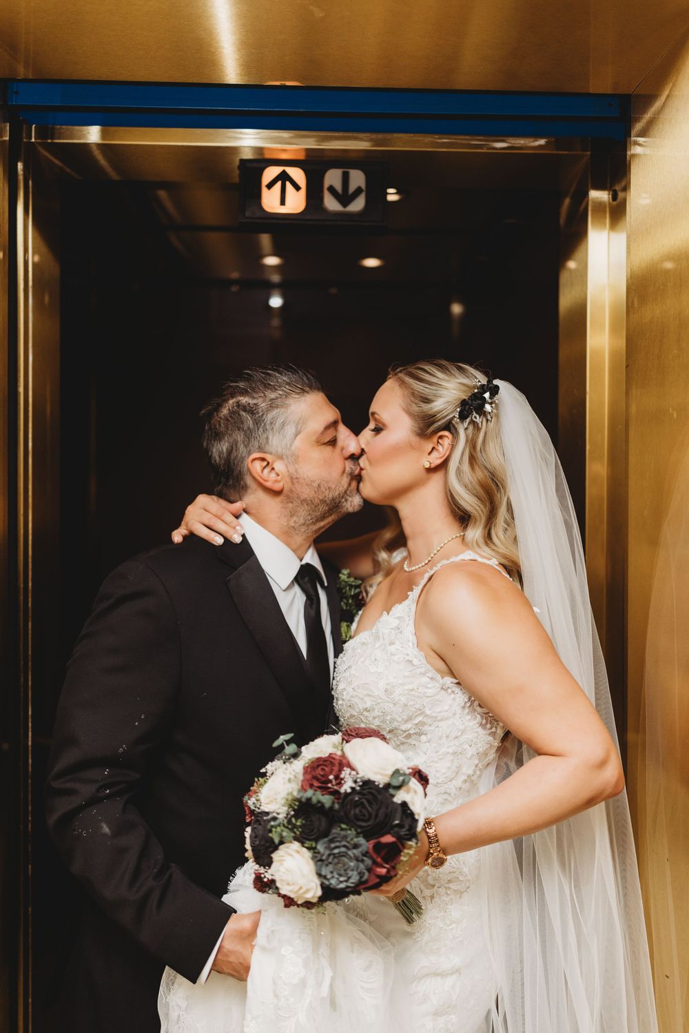 A romantic elevator kiss between newlyweds holding a dark floral bouquet.