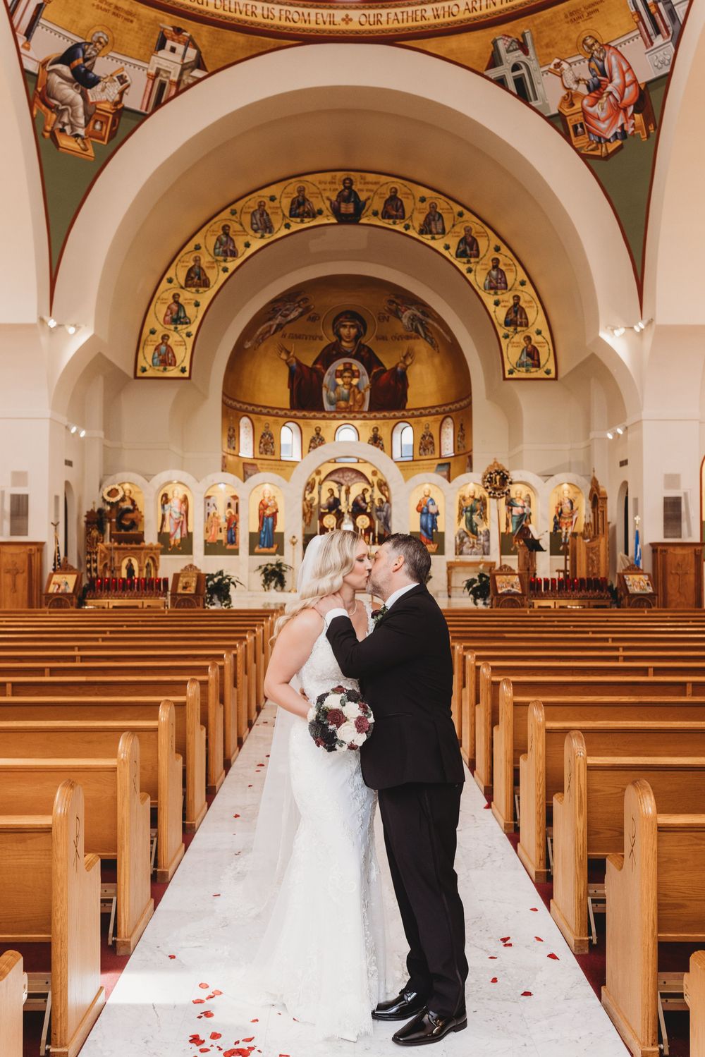 Couple shares intimate wedding ceremony moment in ornate church sanctuary with red rose petals scattered on aisle.