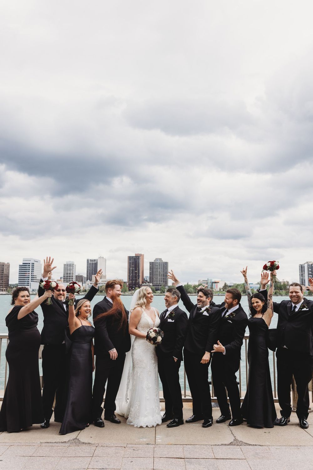 Wedding party celebrates with raised arms against urban waterfront backdrop under cloudy skies.