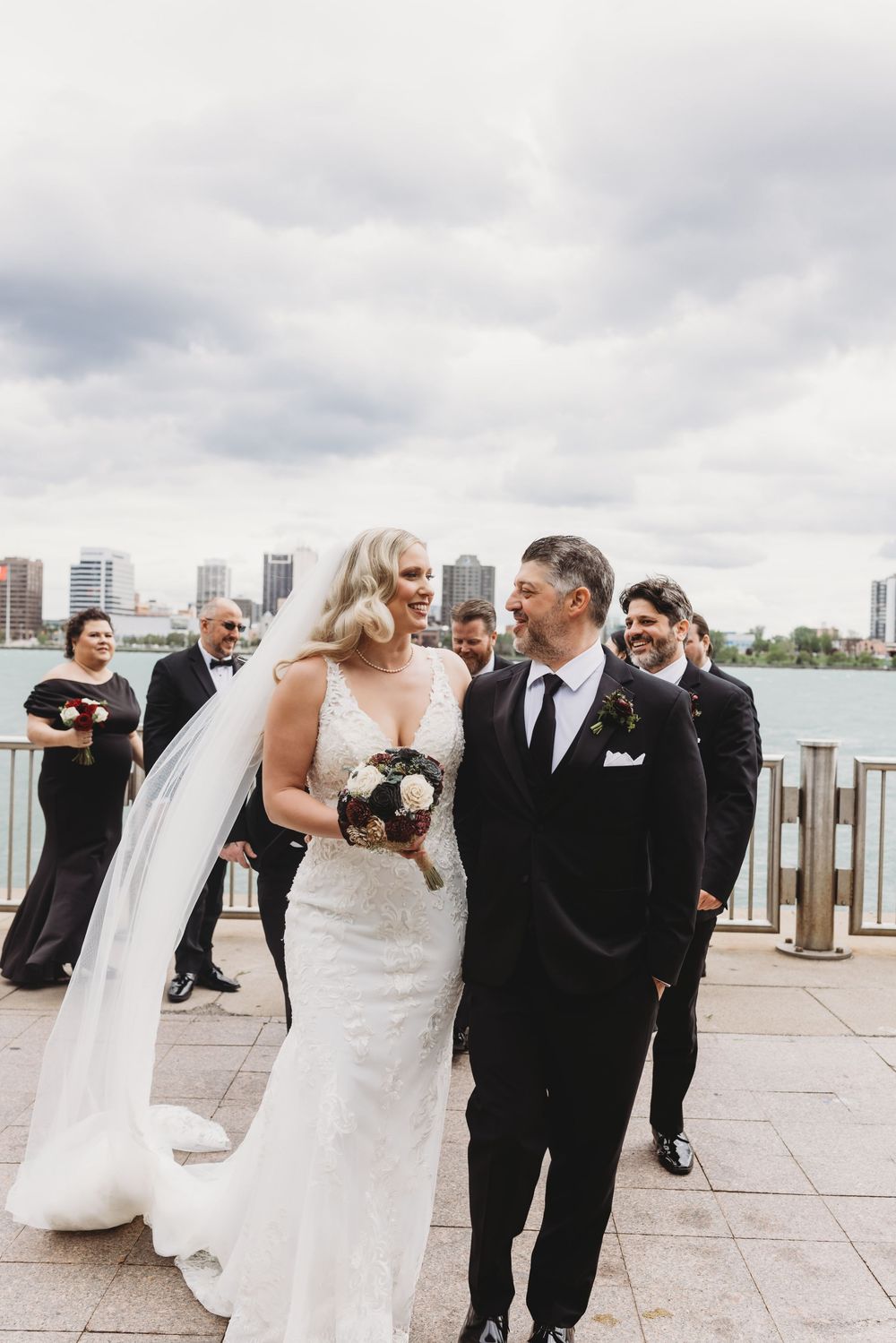 A wedding party poses along a waterfront with a city skyline in the background on an overcast day.