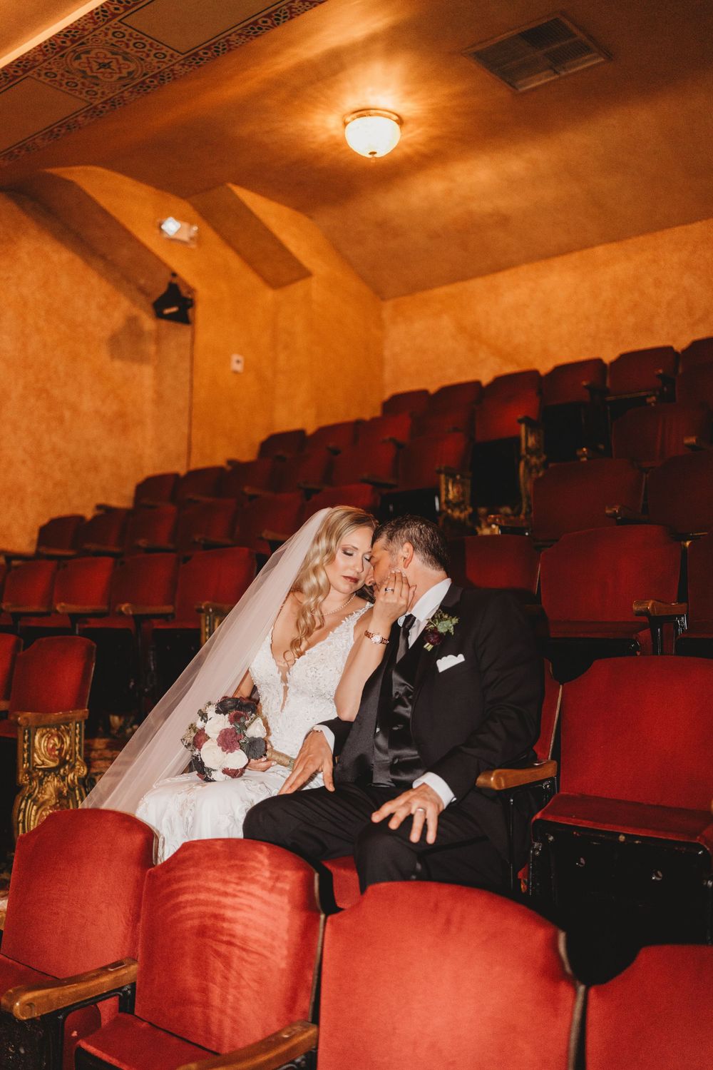 Romantic wedding couple shares intimate moments in vintage theater seats with red velvet upholstery.