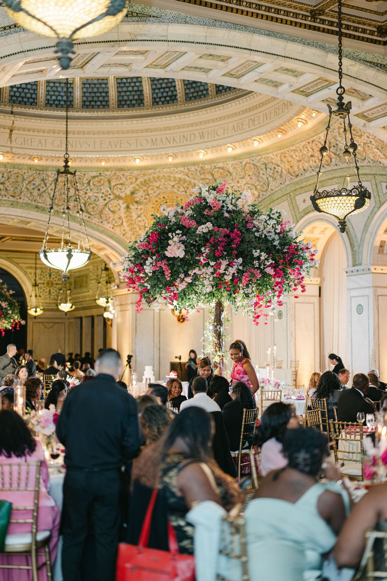 Elegant ballroom wedding reception with ornate ceiling, crystal chandeliers and pink floral arrangements.
