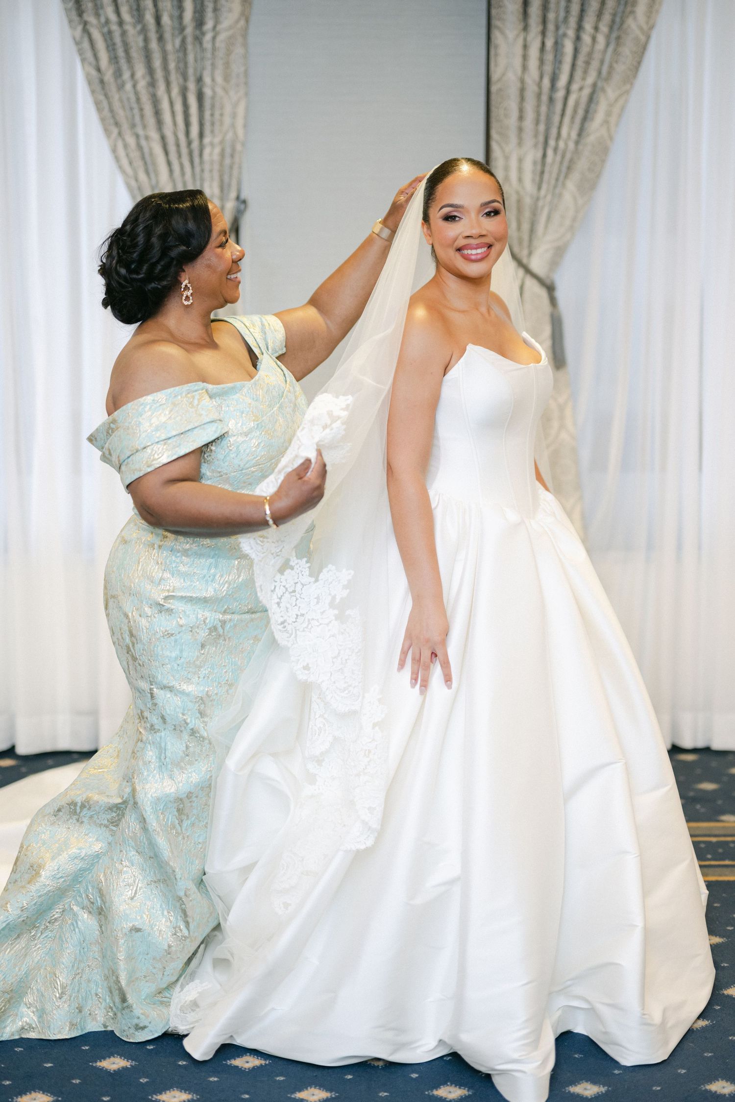 Two women in elegant dresses, one in white and one in light blue, share a joyful moment during a formal event.
