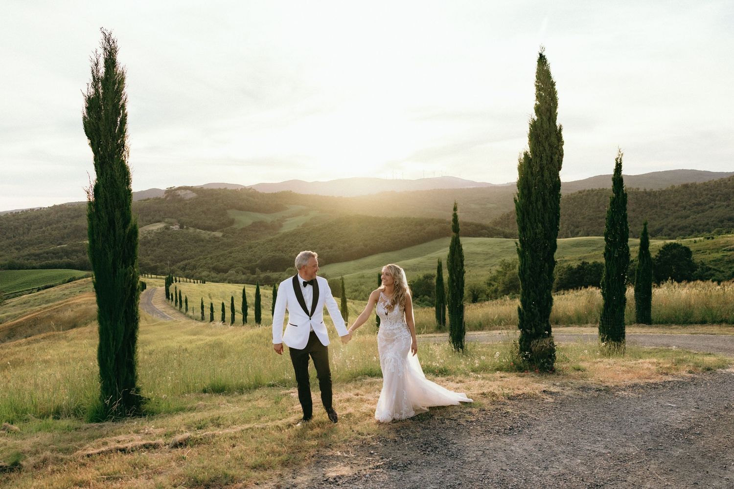 Elegant wedding couple walks along a scenic Tuscan path lined with cypress trees and rolling hills.