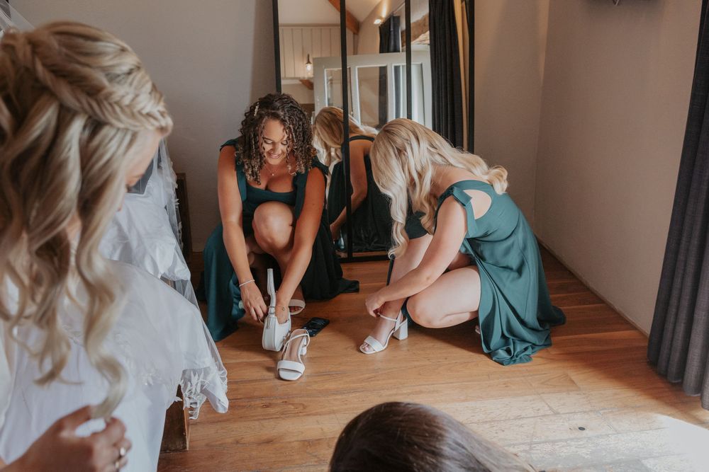 Bridesmaids in green dresses helping bride put on white wedding shoes on wooden floor.