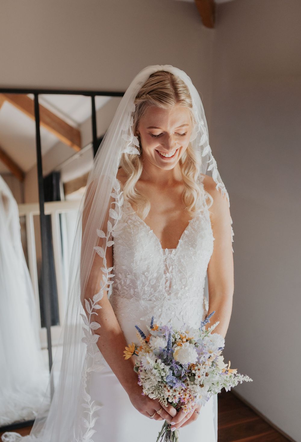 Smiling bride in lace wedding dress and floral veil holding colorful wildflower bouquet.