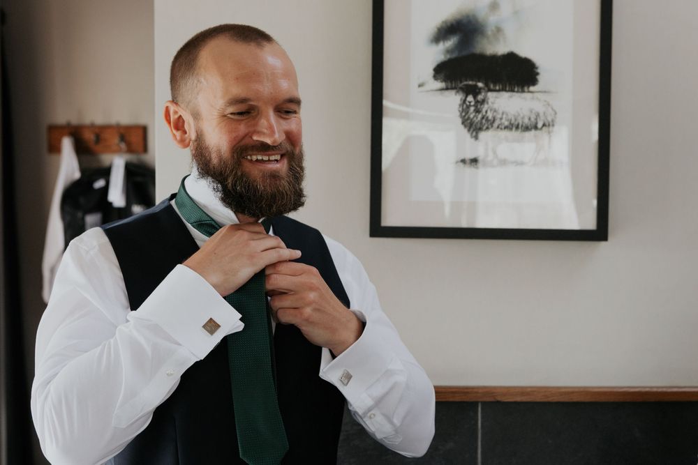 Bearded groom smiling while adjusting green tie in white shirt and grey vest.