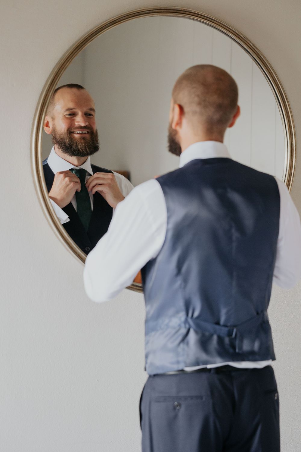 Groom in grey vest adjusting green tie while smiling at reflection in round mirror.