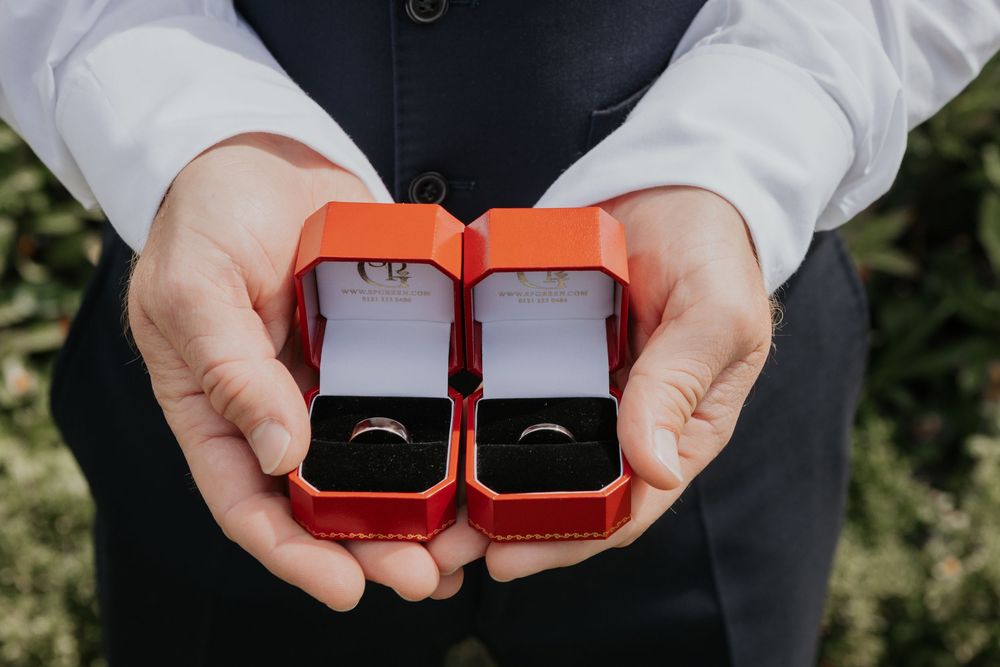 Groom holding two red ring boxes with matching wedding bands on wedding day.