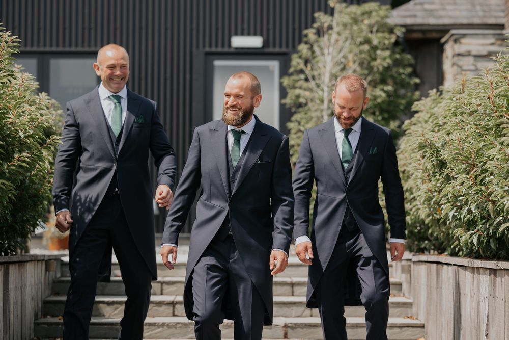 Three groomsmen in grey suits with green ties walking together outdoors.