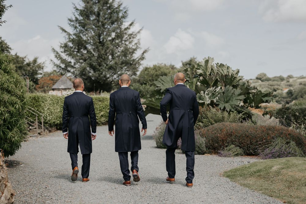 Three groomsmen in dark suits walking away down a garden path at wedding venue.