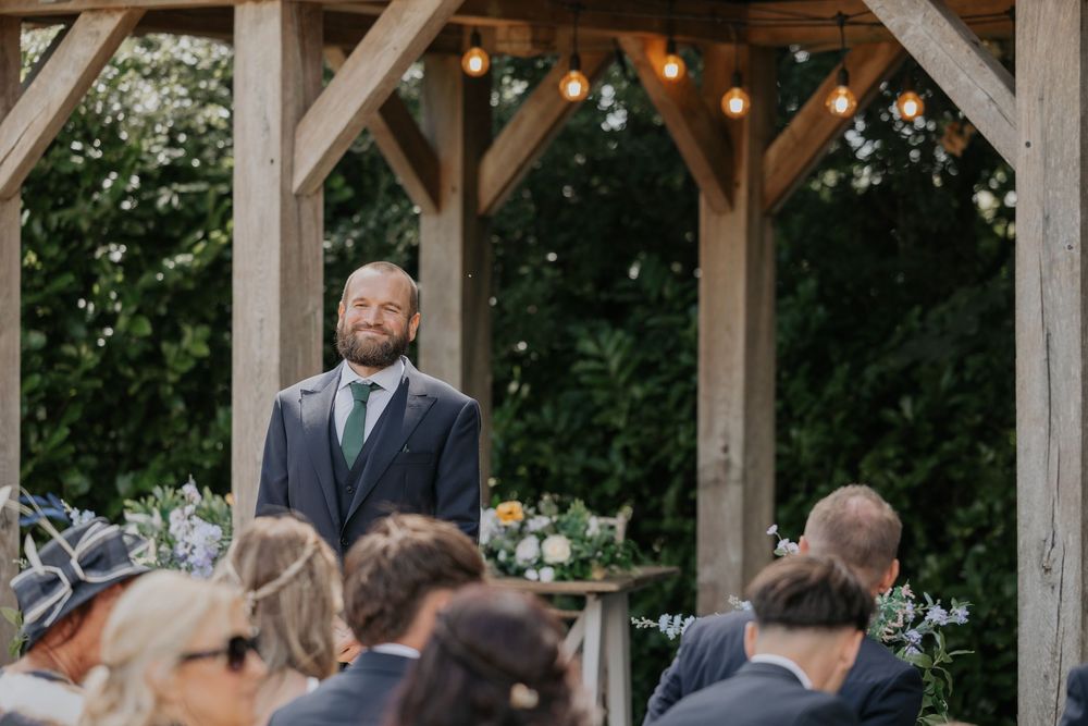 Bearded groom standing at outdoor wooden altar awaiting bride during garden ceremony.
