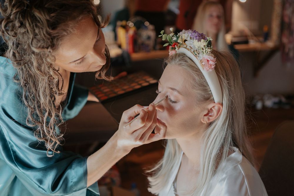 Makeup artist applying finishing touches to smiling bride wearing floral headband during wedding morning preparations.