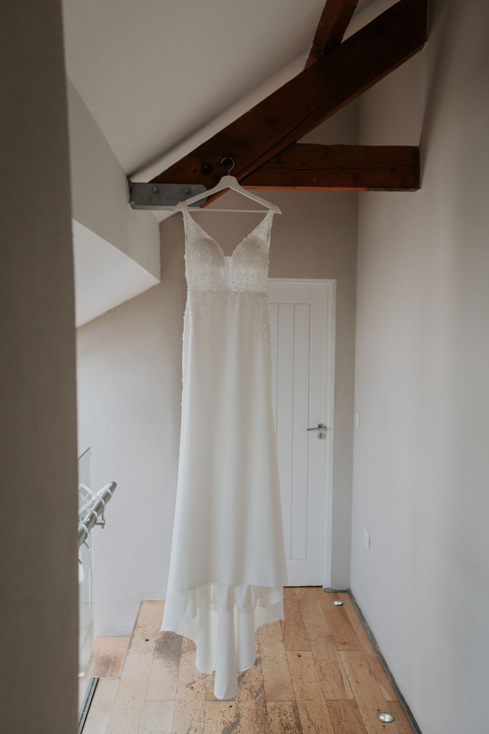 Elegant white wedding dress with lace bodice hanging on hanger in rustic room with wooden beams.