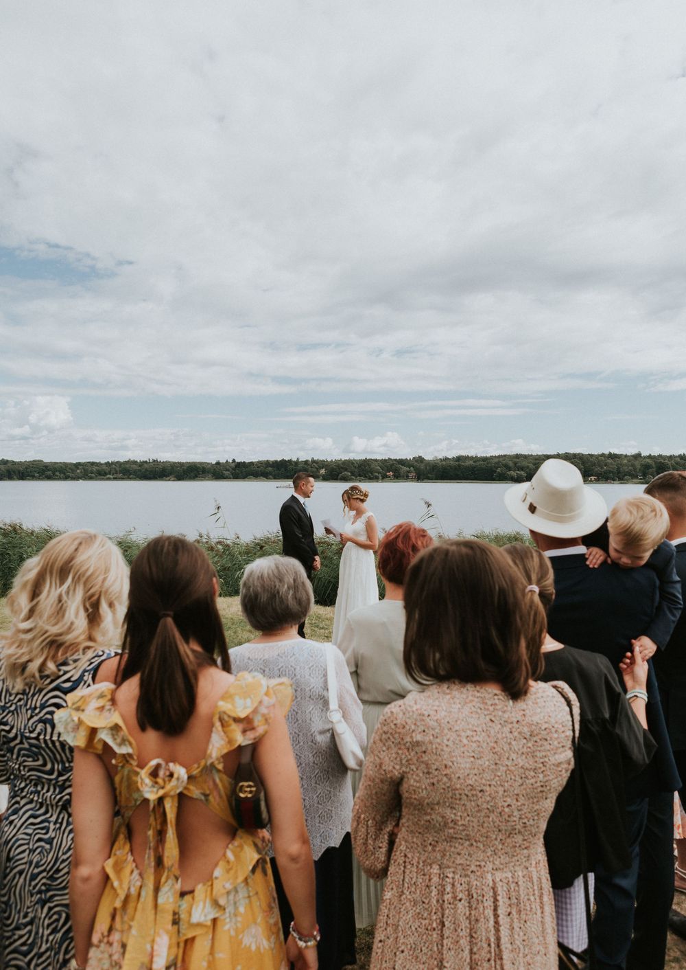 Guests watch an outdoor wedding ceremony by a lake under a cloudy sky.
