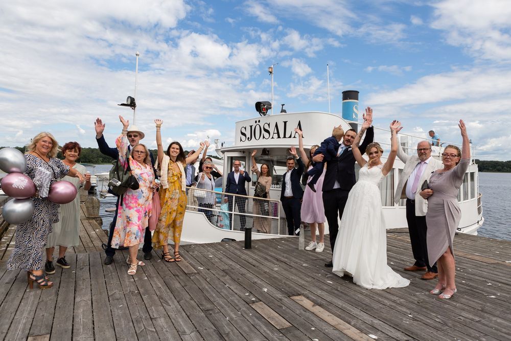 Wedding party celebrates on a dock beside a vintage steamboat on a sunny day.
