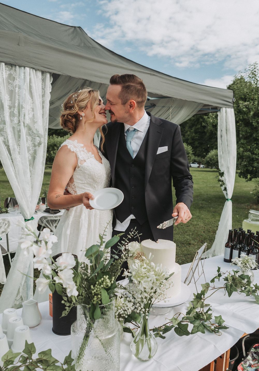 Bride and groom smiling together while cutting wedding cake under a white tent.
