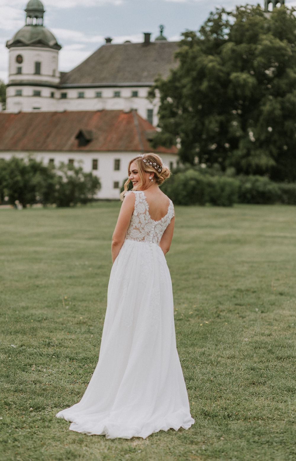 Bride in lace wedding gown with open back standing before a historic castle.