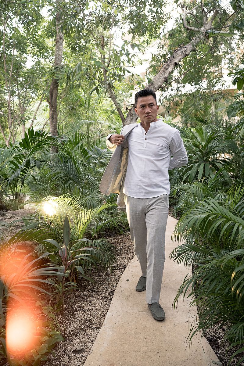 Someone walks along a garden path wearing a white henley shirt and grey pants surrounded by lush tropical plants and palms.