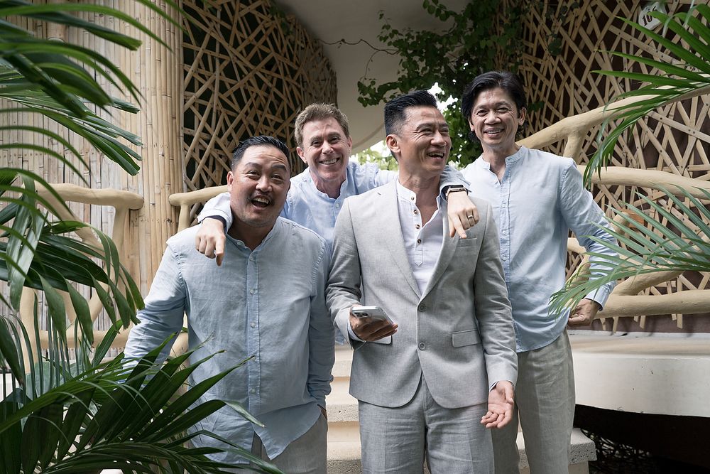 A group wearing white chef coats and uniforms pose together in a tropical setting with palm fronds in the background.