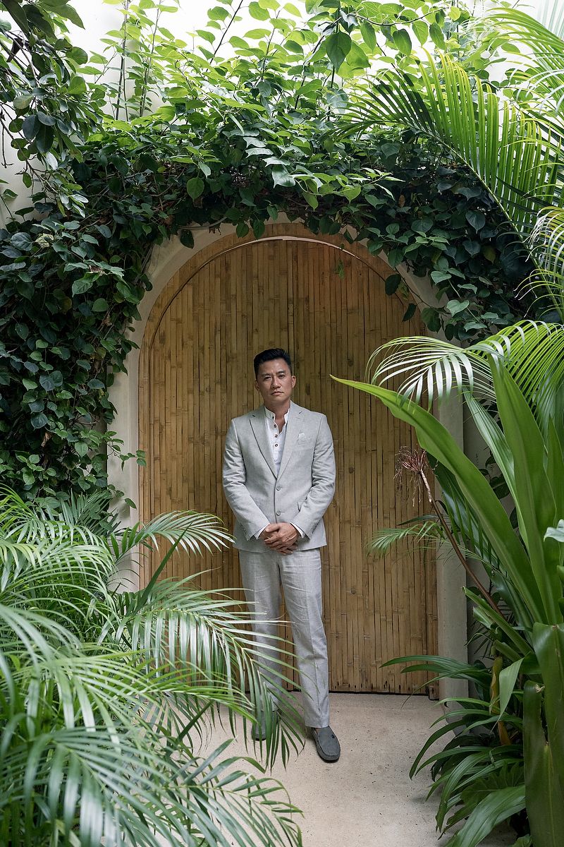 Groom standing beside a wooden arch door at KAN Tulum, surrounded by dense tropical plants in a jungle wedding environment.